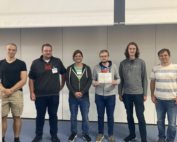 Sechs Personen stehen drinnen und lächeln in die Kamera. Eine Person in der Mitte hält stolz ein Zertifikat der Hochschule Coburg in den Händen. Sie sind leger gekleidet, die schlichten Wände und der schlichte Boden tragen zur entspannten Atmosphäre bei.