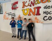 Vier Kinder stehen vor einem bunten Banner mit der Aufschrift „Kinder UNI der Hochschule Coburg“. Sie lächeln fröhlich, halten kleine Karten in den Händen und strecken begeistert den Daumen nach oben. Damit verkörpern sie den Geist dieser Bildungsveranstaltung an der Hochschule Coburg.