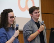 Zwei Personen mit Mikrofonen stehen vor einer Holzwand und einer teilweise rot gestalteten Leinwand in der Hochschule Coburg. Die Person links, mit Locken und Brille, lächelt herzlich neben ihrer kurzhaarigen Begleiterin.