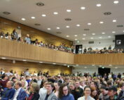Ein großes Publikum füllt einen modernen Hörsaal der Hochschule Coburg. Die Leute sitzen im Erdgeschoss und auf dem Balkon und blicken aufmerksam nach vorne. Der Raum ist mit Holzvertäfelung und Deckenbeleuchtung ausgestattet und schafft eine lebendige, engagierte Atmosphäre.