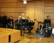 Eine Band der Hochschule Coburg tritt im Innenbereich auf. Die Gruppe besteht aus einem Pianisten, einem Saxophonisten, einem Posaunisten, einem Gitarristen und einem Schlagzeuger. Sie sind dunkel gekleidet und sitzen neben einer Holzwand mit Notenständern, auf denen ihre Notenblätter liegen.