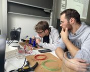 Zwei Personen sind konzentriert und haben Spaß dabei, Elektronik zusammenzubauen. Einer passt Kabel an, während der andere zusieht. Ein Schreibtisch ist vollgestopft mit Werkzeugen, Kabeln und einem Glas und spiegelt die Essenz der Erstsemestertage wider. Im Hintergrund leuchtet eine Präsentation an der Wand, während sie zusammenarbeiten, um zu lernen und zu wachsen.