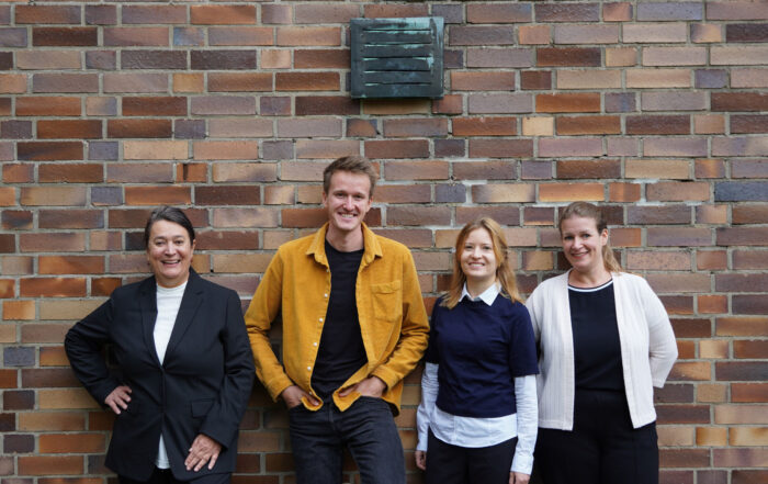 Vier Menschen stehen lächelnd vor einer Backsteinmauer der Hochschule Coburg. Von links nach rechts trägt die erste Person einen schwarzen Blazer, die zweite eine gelbe Jacke, die dritte einen marineblauen Pullover und die vierte eine weiße Strickjacke.
