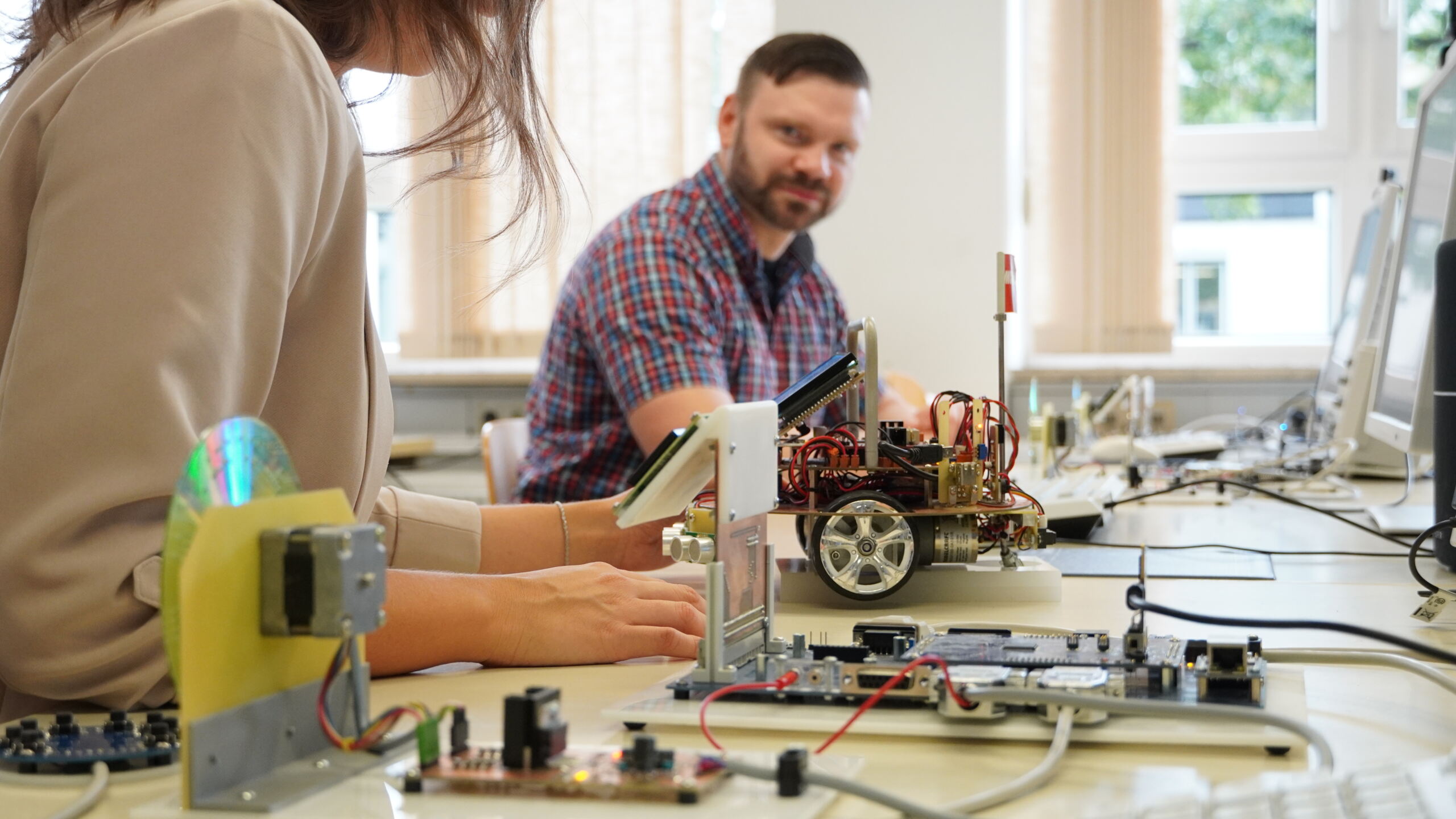 In einem Labor der Hochschule Coburg arbeiten zwei Menschen konzentriert zwischen elektronischen Bauteilen und kleinen Robotern. Leiterplatten und Kabel liegen verstreut auf dem Tisch, während im Hintergrund ein Mann und eine Frau an ihren Projekten arbeiten und Innovationen in Bewegung vorführen.