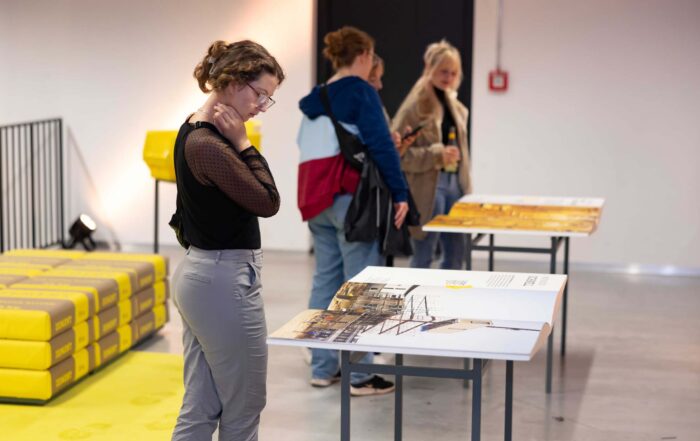 In einer Galerie der Hochschule Coburg betrachten drei Personen Exponate auf Tischen. Eine Person, die eine Brille und ein schwarzes Oberteil trägt, steht näher, während die anderen beiden sich unterhalten. Auf der linken Seite des Raums sind gelbe Taschen gestapelt, die dem Raum einen Farbtupfer verleihen.