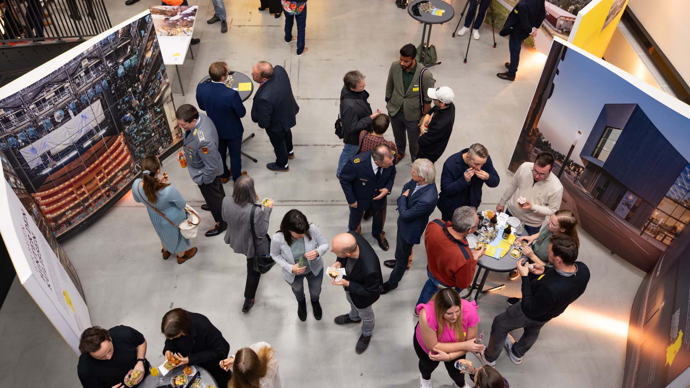 Eine Gruppe von Menschen unterhält sich bei einer Indoor-Veranstaltung, die von großen Anzeigetafeln umgeben ist und wahrscheinlich von der Hochschule Coburg veranstaltet wird. Sie stehen und essen, einige sitzen an hohen Tischen. Die Umgebung ist hell und geräumig, die Teilnehmer sind sowohl in Freizeit- als auch in Business-Kleidung gekleidet.