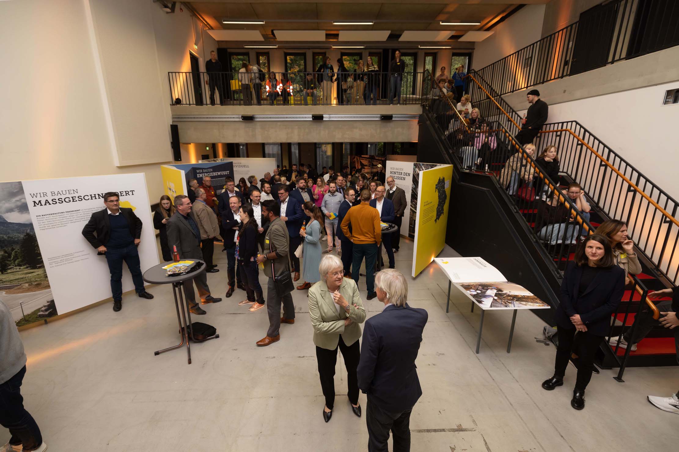 Eine Gruppe versammelt sich in einem geräumigen, modern eingerichteten Innenraum der Hochschule Coburg. Einige stehen neben Schautafeln, während andere sich in kleinen Gruppen unterhalten. Eine Treppe auf der rechten Seite führt in eine obere Etage, wo noch mehr Leute zuschauen.