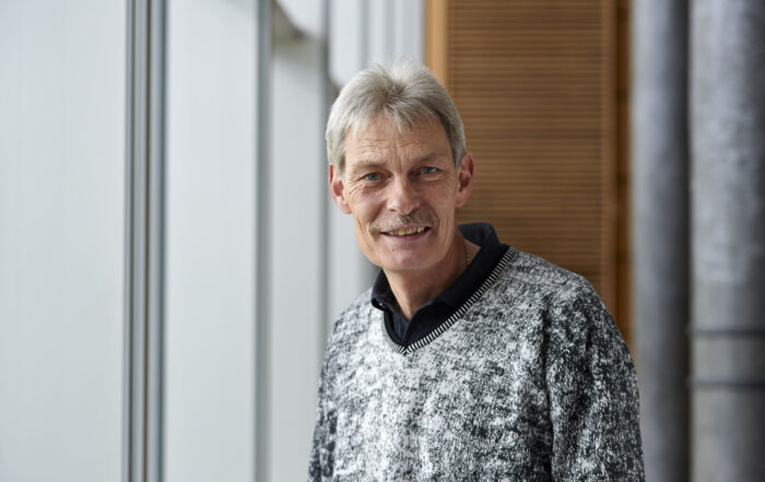 Ein Mann mit kurzen grauen Haaren und einem Schnurrbart steht in einem Gebäude der Hochschule Coburg und lächelt herzlich. Er trägt einen grau-schwarz gemusterten Pullover über einem schwarzen Hemd vor einer Kulisse aus großen Fenstern und Holzpaneelen.