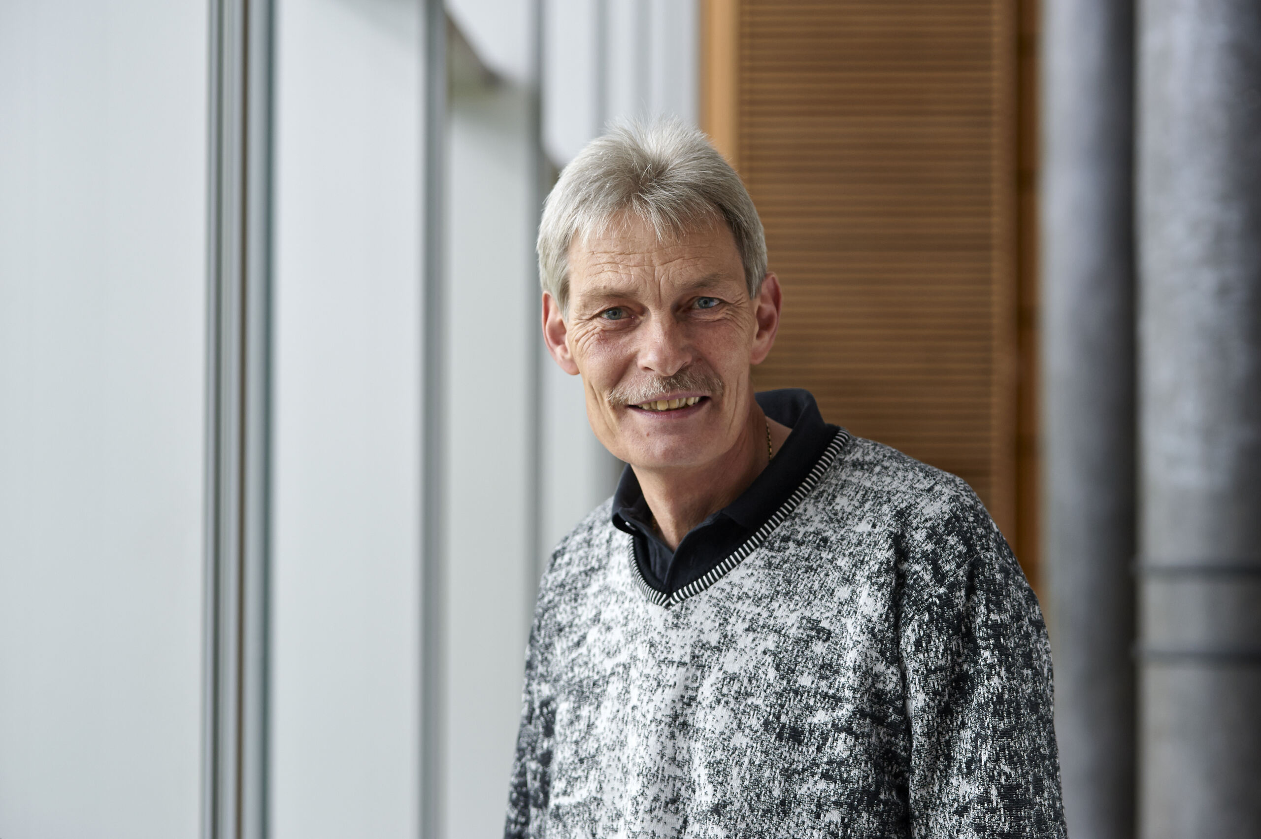 Ein Mann mit kurzen grauen Haaren und einem Schnurrbart steht in einem Gebäude der Hochschule Coburg und lächelt herzlich. Er trägt einen grau-schwarz gemusterten Pullover über einem schwarzen Hemd vor einer Kulisse aus großen Fenstern und Holzpaneelen.