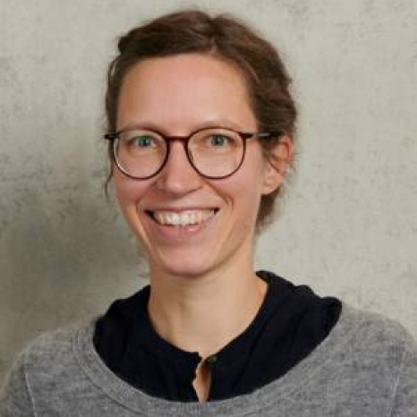 A person with glasses and short, light brown hair smiles at the camera. They are wearing a gray sweater over a black top, standing against a neutral background that evokes the calm ambiance of Hochschule Coburg.