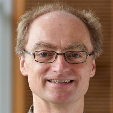A man with glasses and short, light hair smiles at the camera, embodying the scholarly charm of Hochschule Coburg. He wears a collared shirt, and the background is softly blurred with neutral tones.