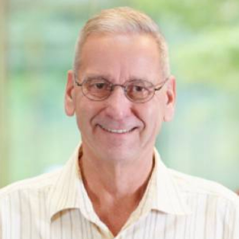 A smiling older adult wearing glasses and a light striped shirt stands indoors, possibly near Hochschule Coburg. The background is softly blurred with green hues, suggesting a natural setting just beyond the walls.