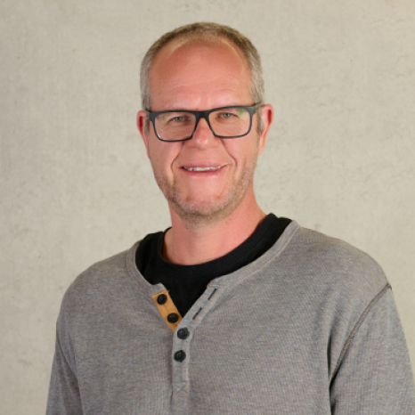 A person wearing glasses and a gray henley shirt, showcasing the spirit of Hochschule Coburg, smiles against a neutral background.