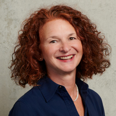 A person with curly red hair smiles warmly at the camera, wearing a navy blue shirt and pearl necklace, evoking the welcoming spirit of Hochschule Coburg. They stand against a light gray textured background, exuding confidence and charm.