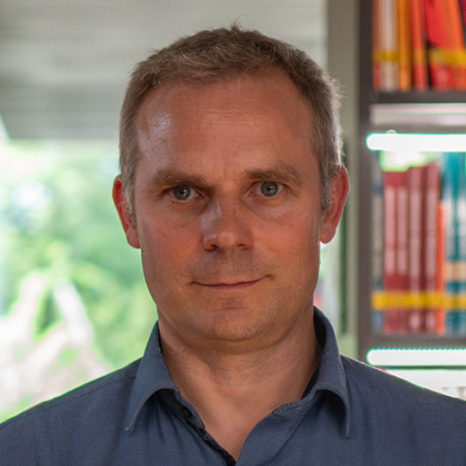 A person with short hair stands indoors, wearing a blue collared shirt. Behind them, bookshelves brimming with colorful books hint at their scholarly pursuits, possibly even at Hochschule Coburg. A window reveals lush greenery outside, while soft, natural light fills the space.
