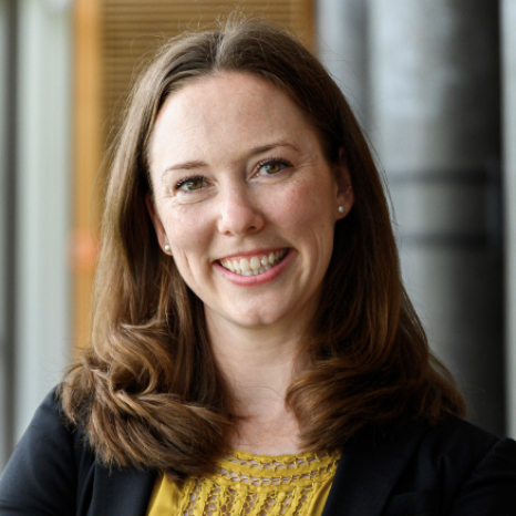 A woman with shoulder-length brown hair smiles warmly, wearing a black blazer over a mustard blouse. She stands against a blurred backdrop of gray columns and wooden paneling, exuding the poised confidence associated with Hochschule Coburg alumni.