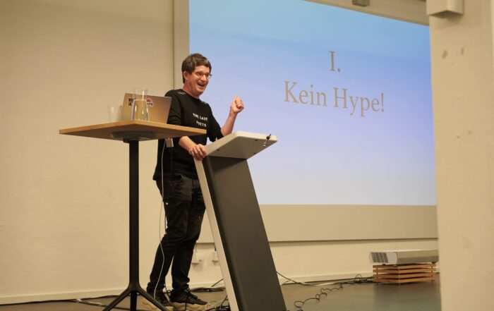 Eine Person steht an einem Rednerpult und hält einen Vortrag. Auf dem Bildschirm hinter ihr erscheint der Text „I. Kein Hype!“. Die Person wirkt beim Sprechen lebhaft und engagiert. Auf dem Rednerpult stehen ein Laptop und ein Glas Wasser.