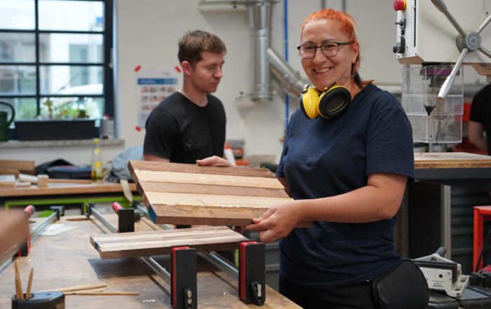 Eine Frau lächelt und hält ein Holzbrett in einer Holzwerkstatt. Sie trägt Sicherheitskopfhörer um den Hals. Im Hintergrund arbeitet ein Mann an einem Tisch mit verschiedenen Werkzeugen und Holzbrettern. Durch das große Fenster fällt natürliches Licht ein.