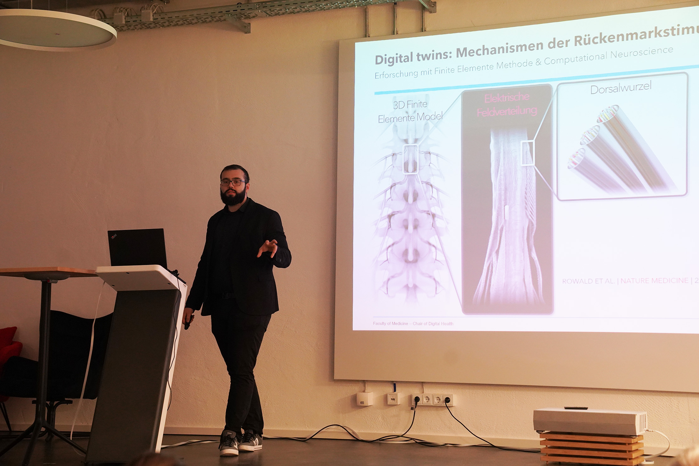 Ein Mann steht neben einem Präsentationsbildschirm, auf dem Bilder und Texte der Wirbelsäule zu sehen sind. Der Raum ist für eine Vorlesung eingerichtet, mit einem Podium und ein paar Stühlen. Die Präsentation handelt von „Digitale Zwillinge: Mechanismen des Rückenmarks“ in einem neurowissenschaftlichen Kontext.