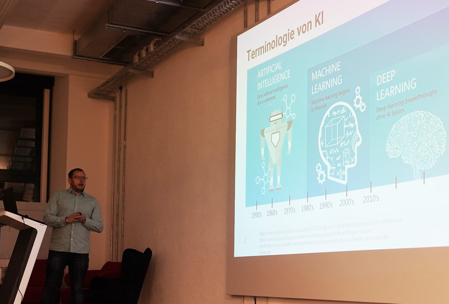 Ein Mann in legerer Kleidung hält einen Vortrag über künstliche Intelligenz, maschinelles Lernen und Deep Learning. Er steht neben einem Rednerpult mit einem Projektor, der in einem modernen Konferenzraum eine Zeitleistengrafik auf einer großen Leinwand anzeigt.
