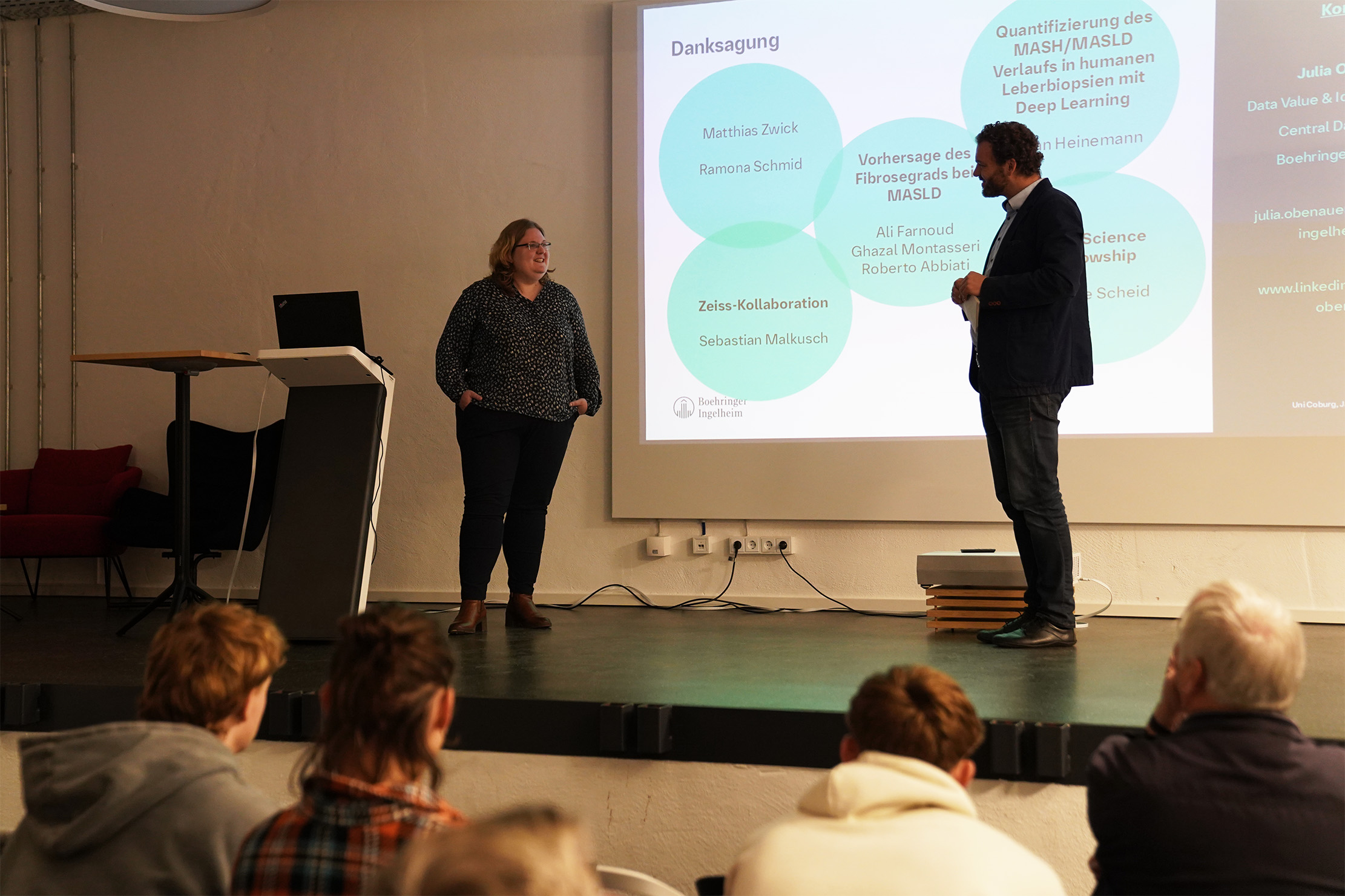 Ein Mann und eine Frau stehen auf einer Bühne und präsentieren ein wissenschaftliches Thema. Die Folie, die hinter ihnen projiziert wird, zeigt miteinander verbundene Kreise mit Namen und Begriffen aus dem Bereich maschinelles Lernen und KI. Vor der Bühne sitzt ein Publikum.
