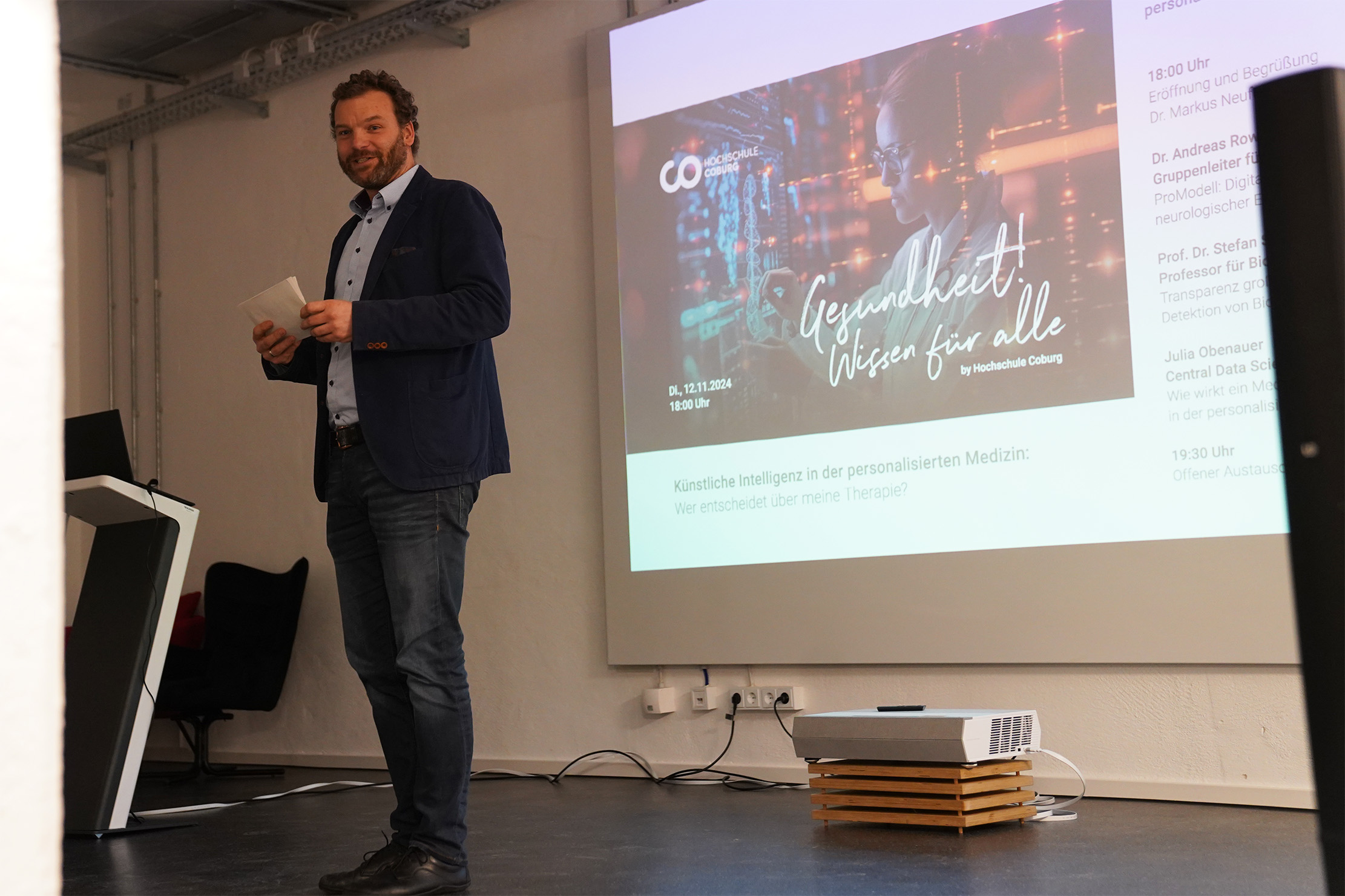 Ein Mann mit einem Papier steht neben einer Projektionsleinwand, auf der eine Präsentation mit dem Titel „Künstliche Intelligenz in der personalisierten Medizin“ mit deutschem Text läuft. Im Hintergrund sind eine weiße Wand und ein Stuhl zu sehen.