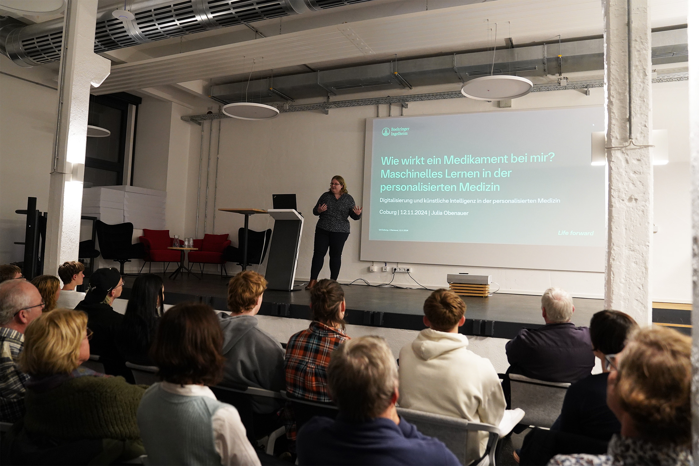 Ein Redner präsentiert eine Folie in einem modernen Hörsaal. Die Folie trägt den Titel „Wie wirkt ein Medikament bei mir? Maschinelles Lernen in der personalisierten Medizin.“ Das Publikum sitzt und verfolgt aufmerksam die Präsentation.
