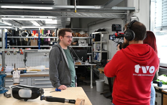 Eine Person in Freizeitkleidung wird in einer Werkstatt von einem zweiköpfigen Kamerateam gefilmt. Im Vordergrund liegt eine Beinprothese auf dem Tisch. Die Werkstatt ist voll mit Werkzeugen, Materialien und Geräten.