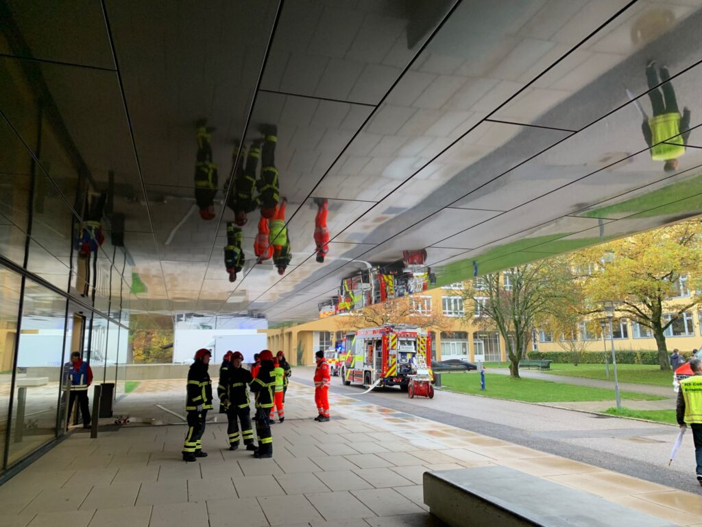Firefighters in uniform stand under a reflective ceiling, with a fire truck parked nearby. The outdoor area is paved, and autumn trees are visible in the background. Reflections of the scene are seen on the ceiling.