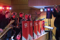 A jazz band performs in a dimly lit space, with musicians playing saxophones, trumpets, and trombones. A conductor stands to the right, leading the ensemble. Red banners with white logos and text are displayed in front of the musicians. at Hochschule Coburg