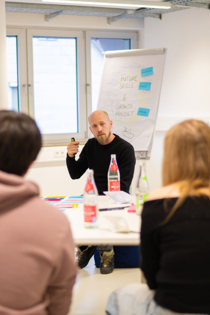 Ein Mann sitzt und gestikuliert, während er zu einer Gruppe um einen Tisch in einem hell erleuchteten Raum spricht. Die Gruppe ist aufmerksam, auf dem Tisch stehen Notizbücher und Getränke. Im Hintergrund ist ein Flipchart mit Notizen darauf. at Hochschule Coburg