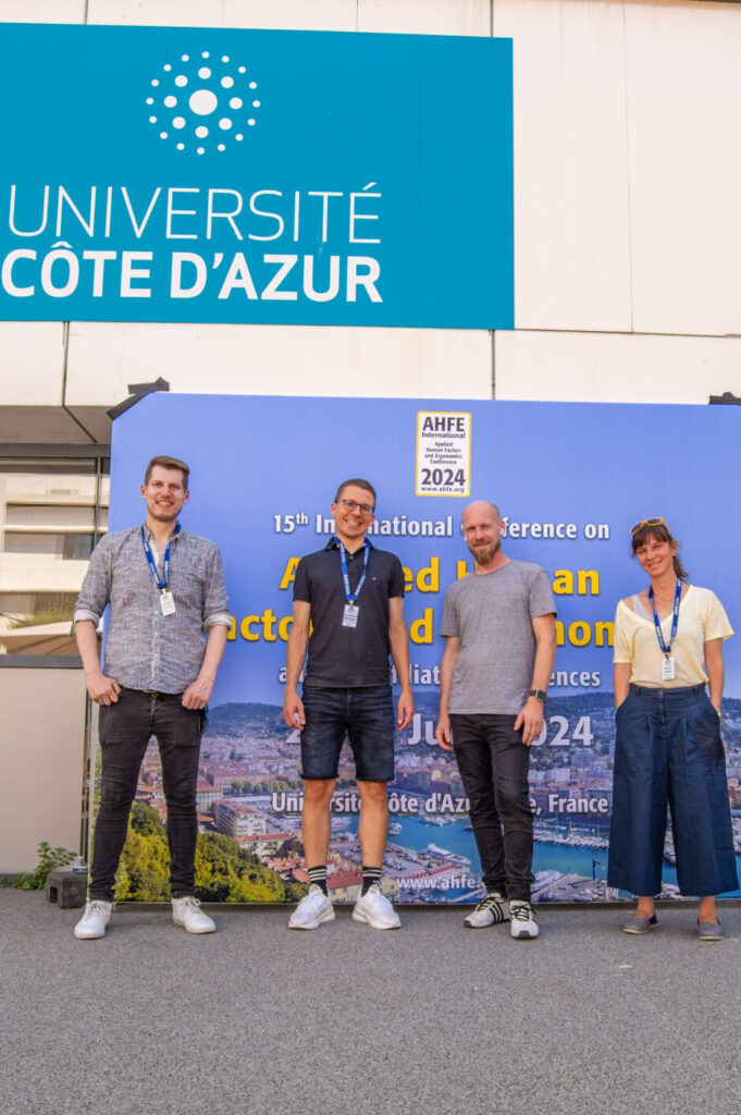 Vier Personen stehen lächelnd vor einem blauen Konferenzhintergrund vor der Université Côte d’Azur. Der Hintergrund zeigt einen Text über eine AMSE-Veranstaltung. In der Nähe befinden sich eine Pflanze und ein kleines Podium. Auf der Gebäudefassade sind Logos und Texte zu sehen. at Hochschule Coburg