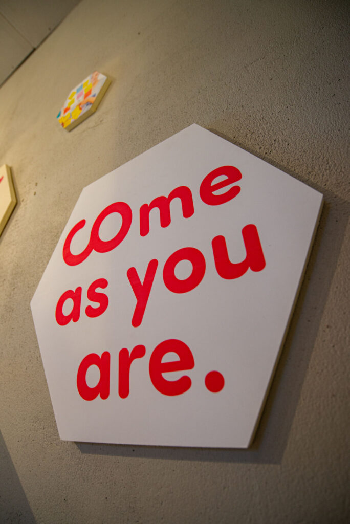 Sechseckiges Schild an einer strukturierten Wand mit dem Satz „Come as you are“ in roten Kleinbuchstaben. Im Hintergrund sind teilweise weitere bunte Dekorationen sichtbar. at Hochschule Coburg