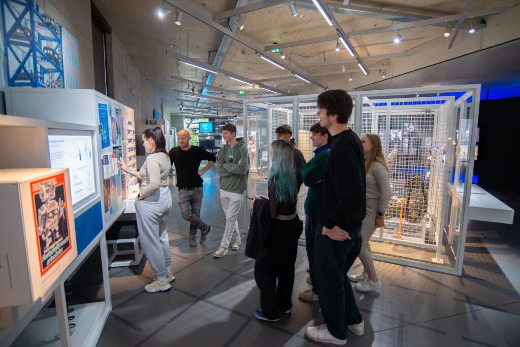 Eine Gruppe von Menschen steht in einer modernen Museumsausstellung. Sie betrachten interaktive Displays an den Wänden. Der Raum ist modern gestaltet, mit schrägen Decken und Beleuchtung. Im Hintergrund ist eine käfigartige Struktur zu sehen. at Hochschule Coburg