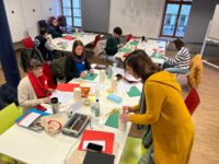 People are seated around tables in a classroom, engaged in various activities with art supplies and papers. A person in a yellow sweater is standing, working on a project. The room has windows and a wooden floor. at Hochschule Coburg