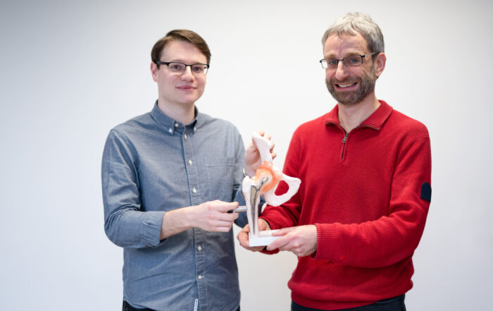 Zwei Männer lächeln und halten ein 3D-Modell eines Beckengelenks in der Hand. Der Mann links im grauen Hemd und der Mann rechts im roten Pullover präsentieren ihre Forschung zu Hüftprothesen vor einem schlichten weißen Hintergrund. at Hochschule Coburg