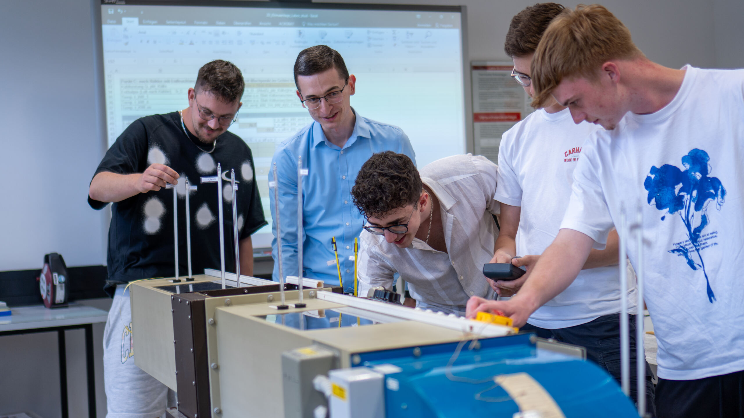 Fünf Personen haben sich um den Aufbau eines physikalischen Experiments versammelt. Sie konzentrieren sich auf eine Tischvorrichtung mit Drähten und Gewichten. Im Hintergrund ist ein digitaler Bildschirm zu sehen, auf dem Daten angezeigt werden. at Hochschule Coburg