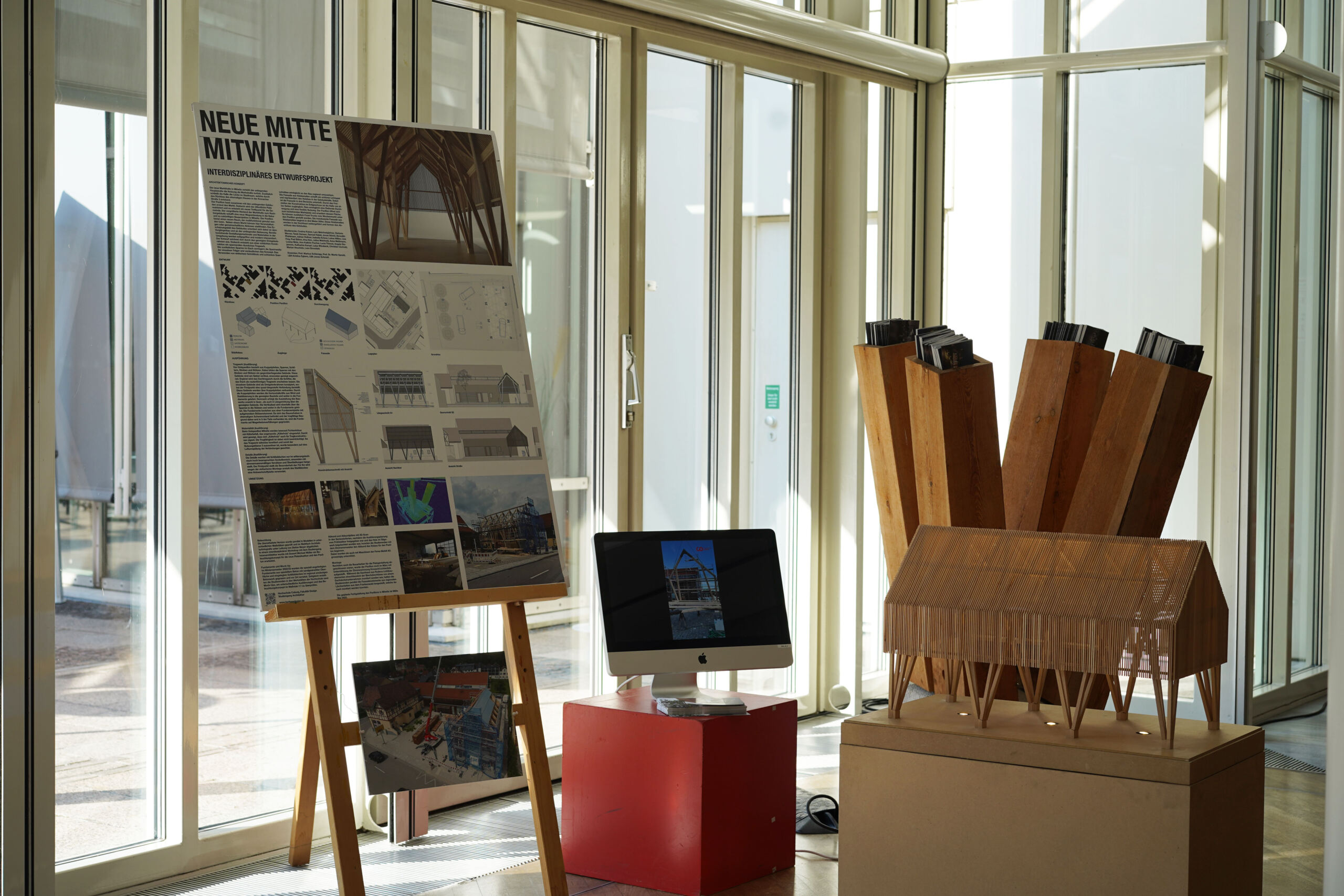 Ein Ausstellungsaufbau mit einer großen Plakatwand mit Architekturskizzen und -fotos, ergänzt durch einen Computerbildschirm auf einem roten Ständer und ein hölzernes Architekturmodell auf einem Podest. Das Ganze befindet sich in einem sonnendurchfluteten Raum mit Glaswänden, der sich perfekt für interaktive Rundgänge eignet. at Hochschule Coburg