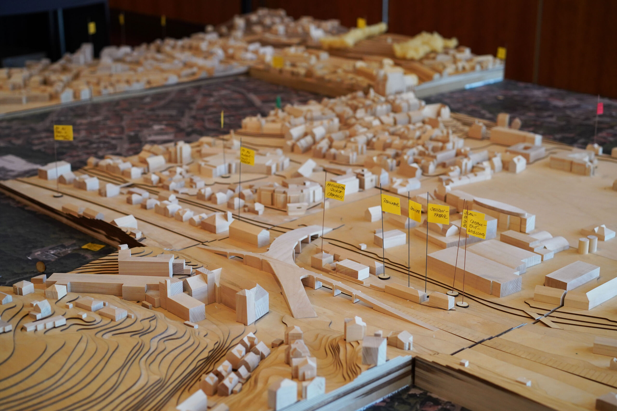 Ein detailliertes Architekturmodell aus Holz, das eine Stadtlandschaft mit Gebäuden, Straßen und topografischen Elementen darstellt. Gelbe Flaggen mit Beschriftungen markieren bestimmte Bereiche oder Strukturen und laden zu Erkenntnissen in Workshops ein. Der Schwerpunkt liegt auf Stadtplanung und -entwicklung. at Hochschule Coburg