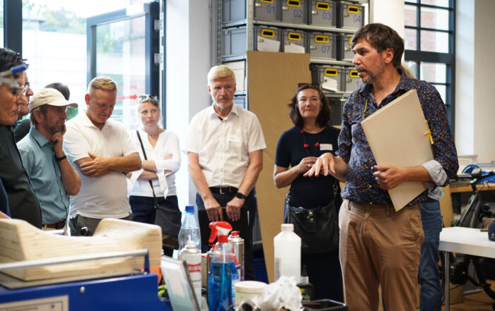 Ein Mann mit einer Tafel steht in einer Werkstatt und spricht zu einer Gruppe Erwachsener, die sich zum Denkmalschutz-Kongress versammelt haben. Die Gruppe, darunter Männer und Frauen mit verschränkten Armen, konzentriert sich auf den Redner. Im Hintergrund stehen Regale und Tische voller verschiedener Gegenstände. at Hochschule Coburg