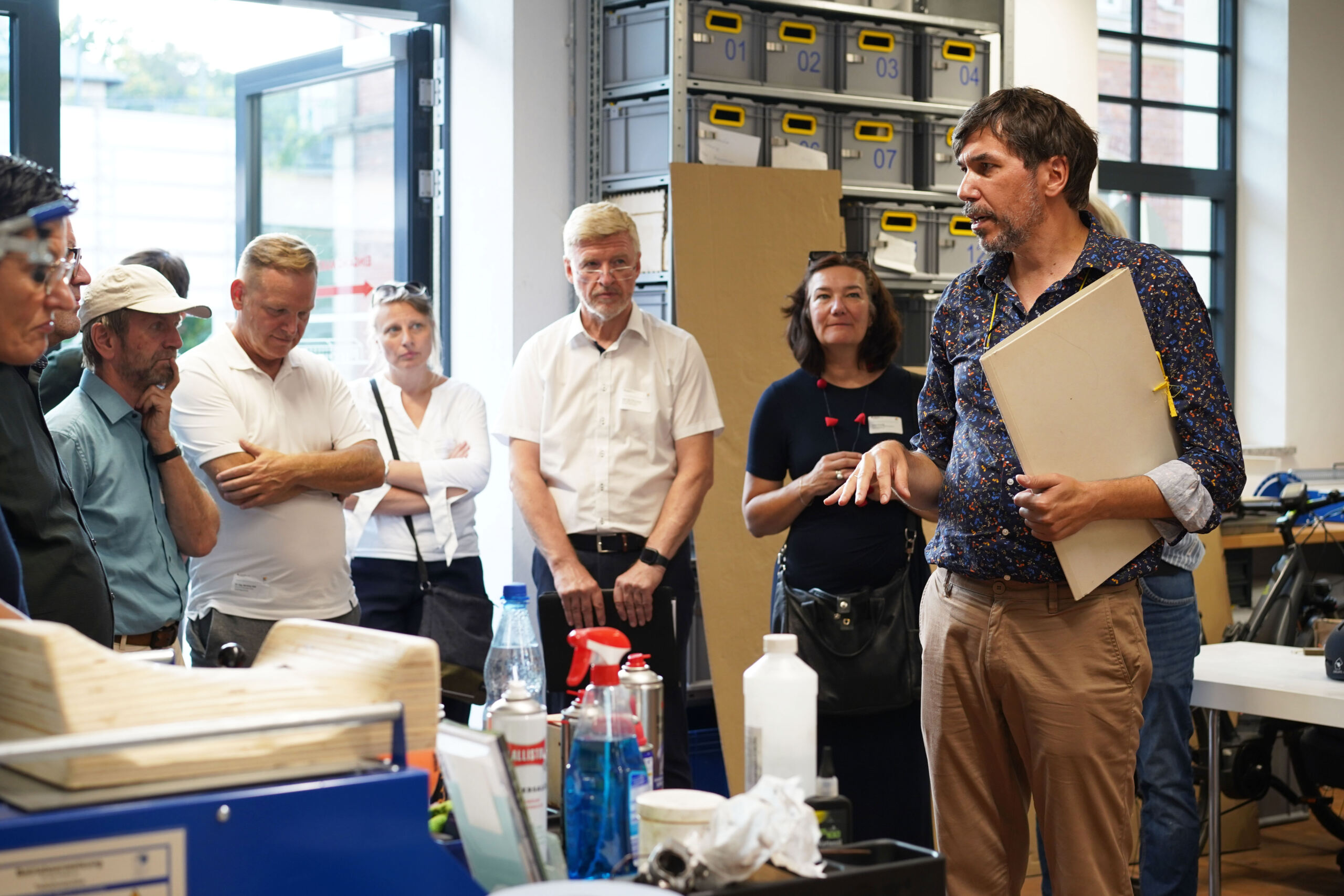 Ein Mann mit einer Tafel steht in einer Werkstatt und spricht zu einer Gruppe Erwachsener, die sich zum Denkmalschutz-Kongress versammelt haben. Die Gruppe, darunter Männer und Frauen mit verschränkten Armen, konzentriert sich auf den Redner. Im Hintergrund stehen Regale und Tische voller verschiedener Gegenstände. at Hochschule Coburg