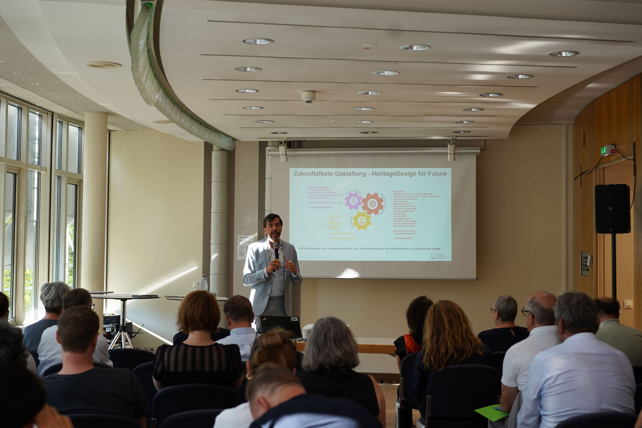 Ein Redner steht vor einem Raum beim Denkmalschutz-Kongress und hält einen Vortrag mit dem Titel „Zukunftsfähige Gestaltung – Heritage Design for Future“. Hinter ihm zeigt eine Projektionsleinwand bunte Zahnräder und Text. Die Teilnehmer sitzen, beteiligen sich eifrig an den Workshops und blicken dem Redner zu. at Hochschule Coburg