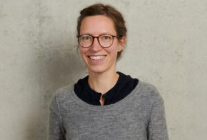 Smiling person with glasses, wearing a gray sweater and dark shirt, stands against a plain wall. at Hochschule Coburg
