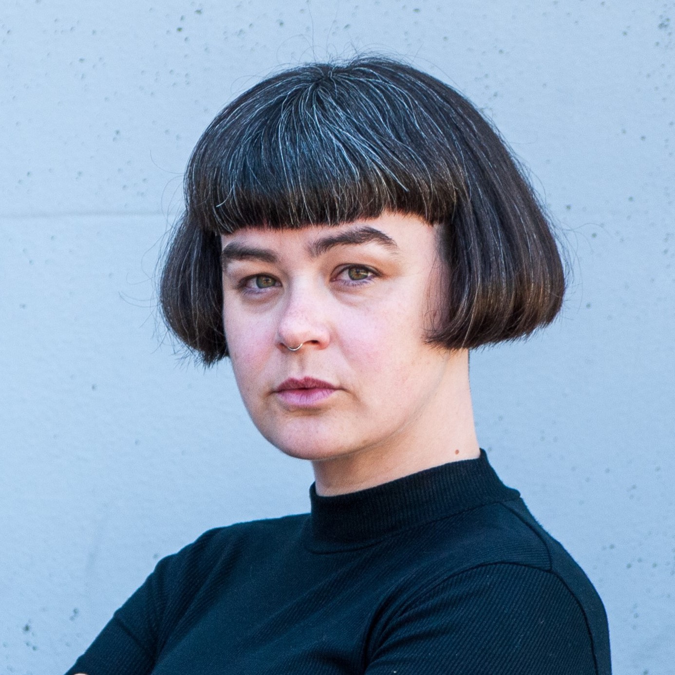 A person with short, straight brown hair featuring gray streaks and blunt bangs, wearing a black turtleneck, stands against a light gray wall, looking toward the camera with a neutral expression. at Hochschule Coburg