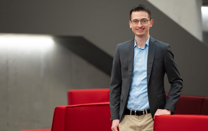 Eine Person in Anzug und hellblauem Hemd steht lächelnd neben roten Möbeln in einem modernen Innenraum mit grauen Wänden und geometrischen Designelementen. at Hochschule Coburg