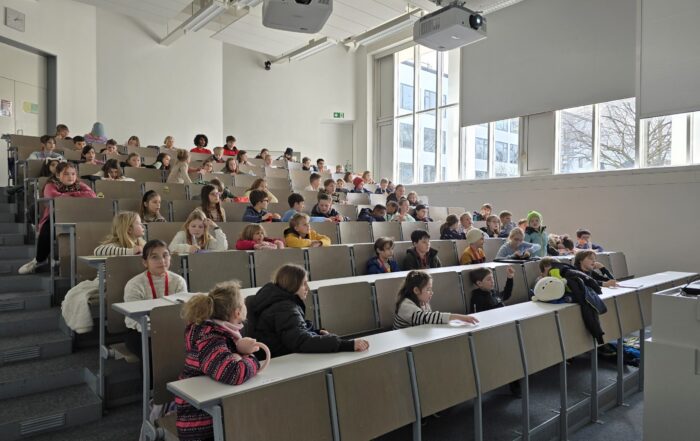 Ein Hörsaal voller Kinder, die in Reihen sitzen und der Kinder-Uni vorne zuhören. Der Raum ist hell, mit großen Fenstern auf der einen Seite und weißen Wänden. Die Kinder sind leger gekleidet und saugen bei dieser besonderen Jubiläumsveranstaltung eifrig Wissen auf. at Hochschule Coburg