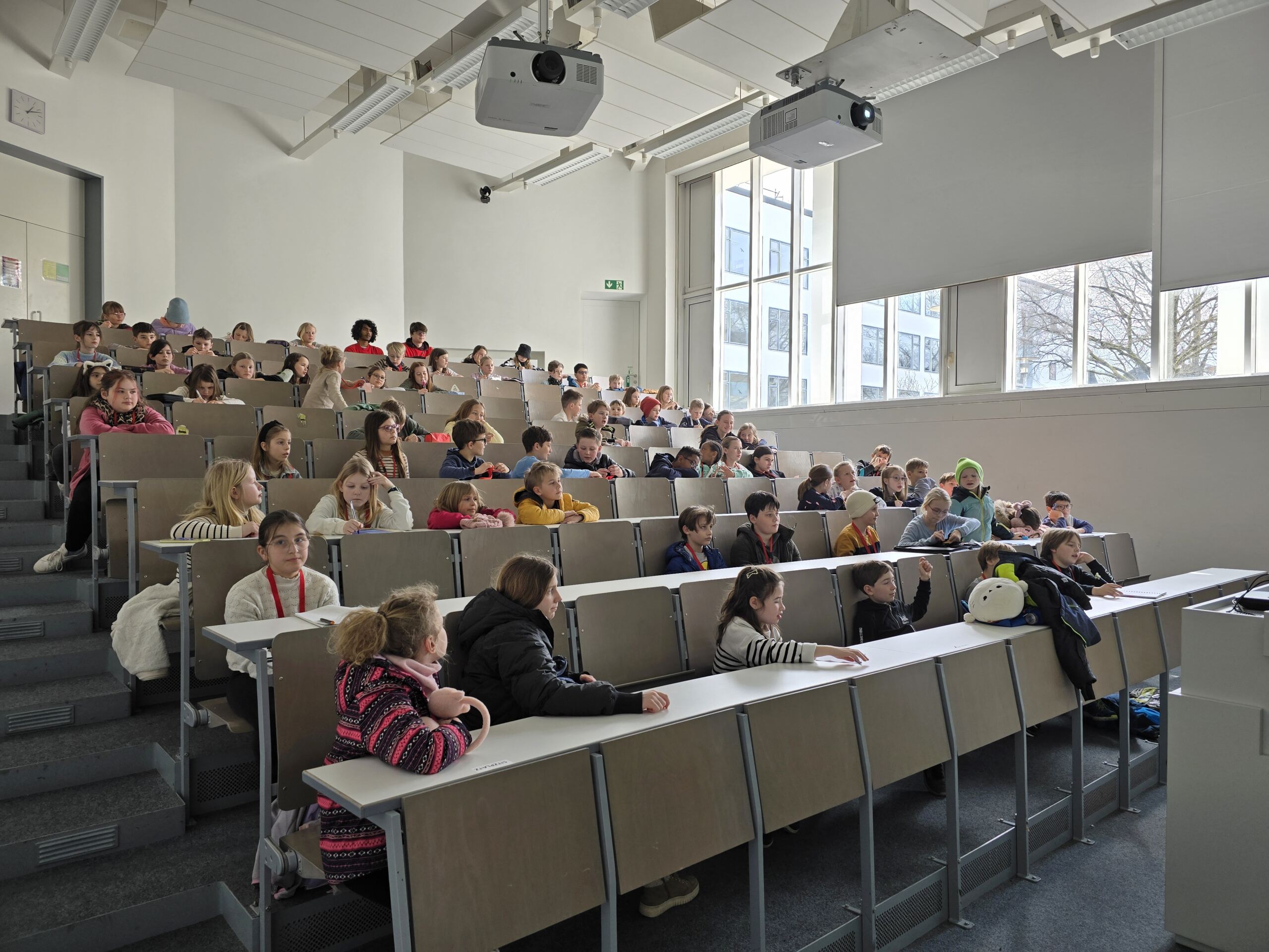 Ein Hörsaal voller Kinder, die in Reihen sitzen und der Kinder-Uni vorne zuhören. Der Raum ist hell, mit großen Fenstern auf der einen Seite und weißen Wänden. Die Kinder sind leger gekleidet und saugen bei dieser besonderen Jubiläumsveranstaltung eifrig Wissen auf. at Hochschule Coburg