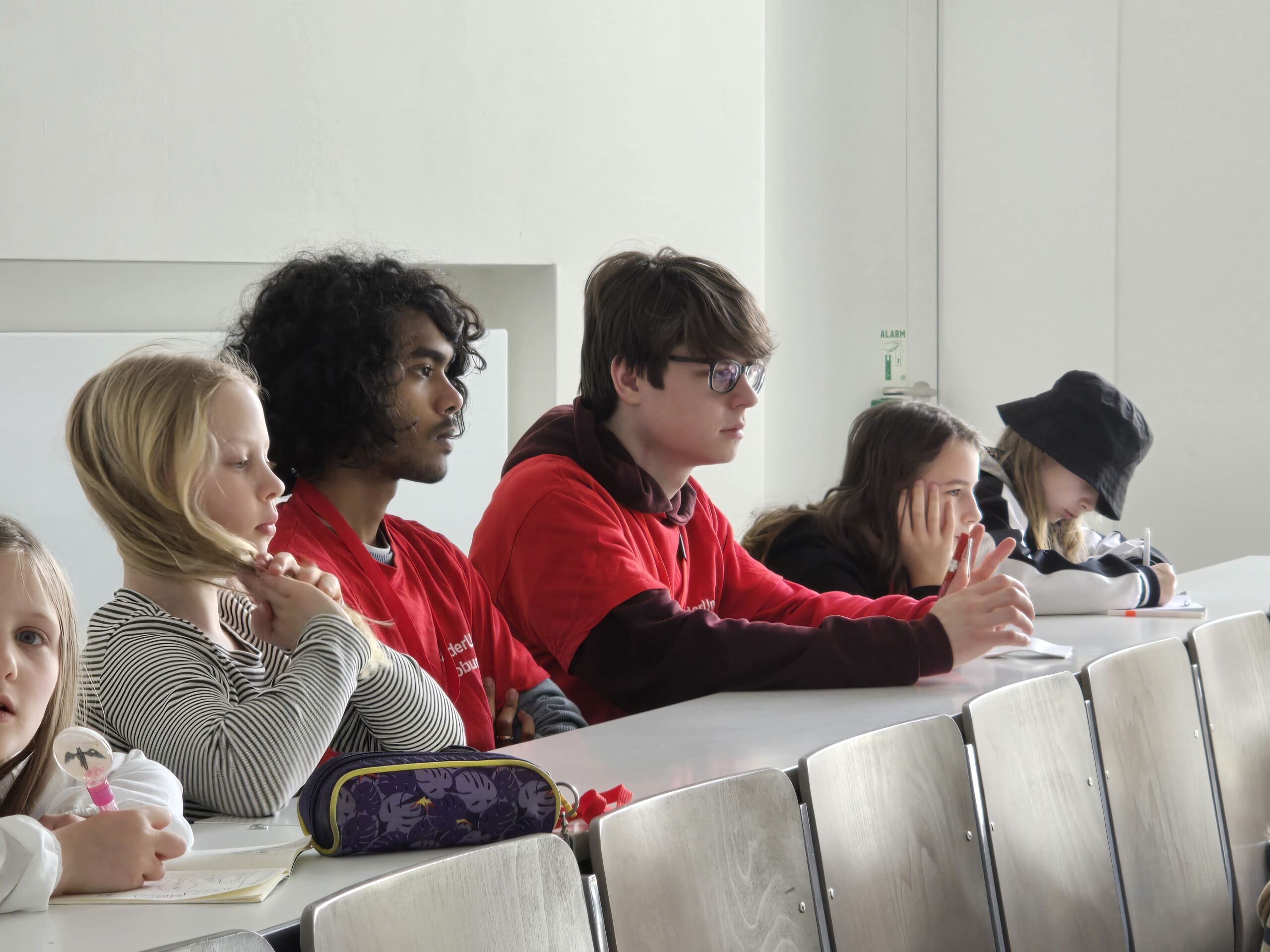 Fünf Studierende sitzen in einem Hörsaal und lauschen aufmerksam einer Kinder-Uni-Vorlesung. Zwei tragen passende rote T-Shirts, einer macht sich Notizen. Der Raum ist hell, und die Studierenden wirken konzentriert, während im Hintergrund Tageslicht durch das Fenster fällt. at Hochschule Coburg