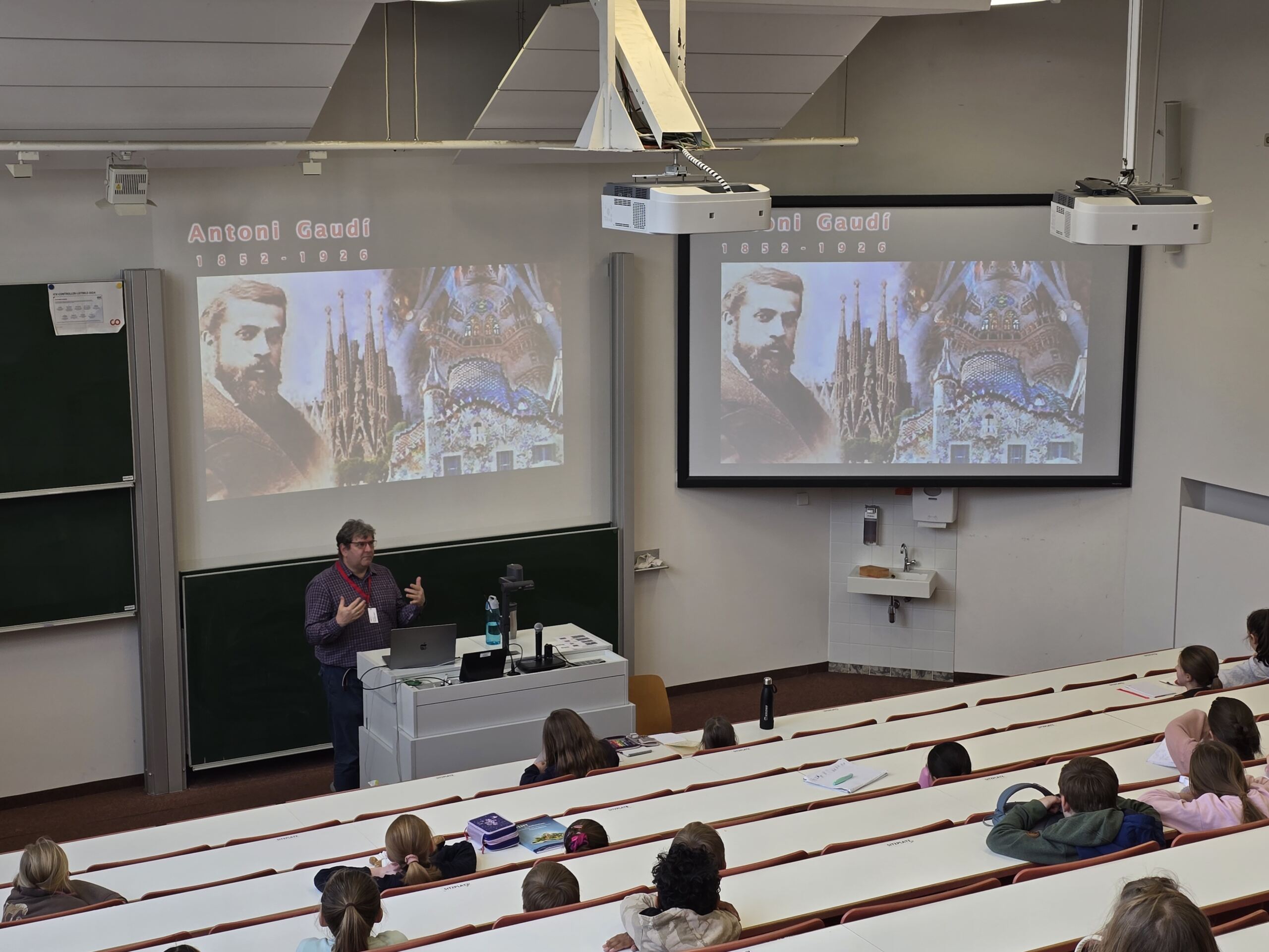 Ein Dozent vor einem Kinder-Uni-Klassenzimmer präsentiert den in Reihen sitzenden Studierenden. Zwei Bildschirme zeigen Antoni Gaudí und die Sagrada Família. Der Raum ist gut beleuchtet und verfügt über einen Overheadprojektor und ein Roboterhund-Modell, was dem Unterrichtsraum einen modernen Touch verleiht. at Hochschule Coburg