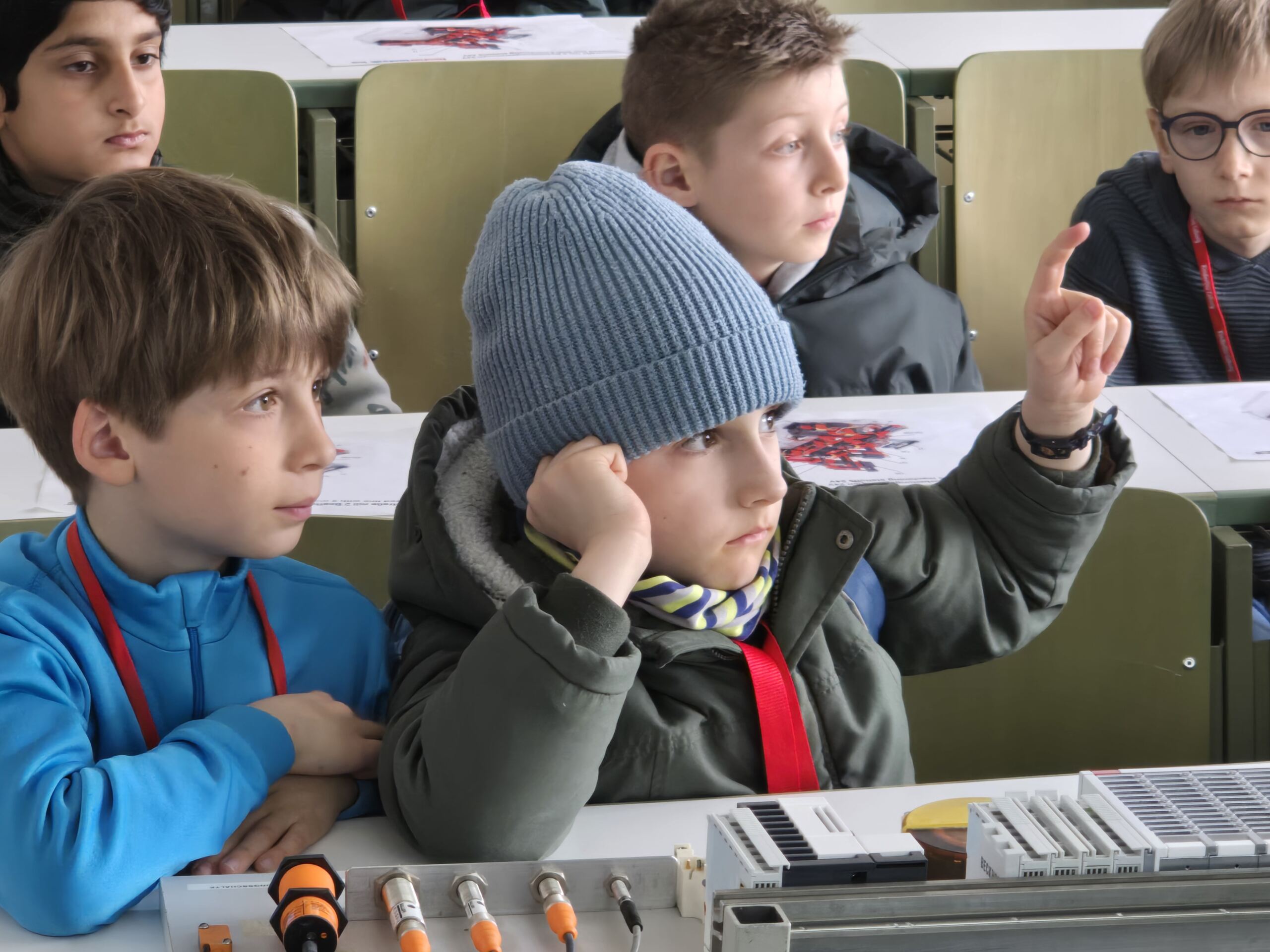 Kinder sitzen an ihren Tischen und nehmen an einer Kinder-Uni-Aktivität teil. Ein Junge mit blauer Mütze hebt interessiert die Hand. Auf ihren Tischen liegen Materialien und Diagramme, alle tragen Schlüsselbänder. at Hochschule Coburg